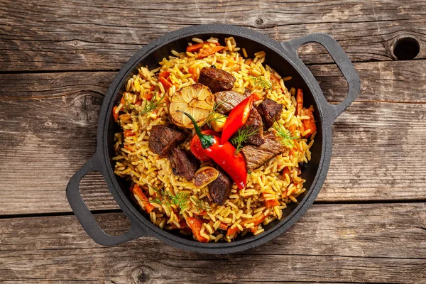 O conceito de cozinha oriental. Pilaf uzbeque nacional com carne em uma frigideira de ferro fundido, em uma mesa de madeira. imagem de fundo. vista superior, espaço de cópia, leigos planos — Fotografia de Stock