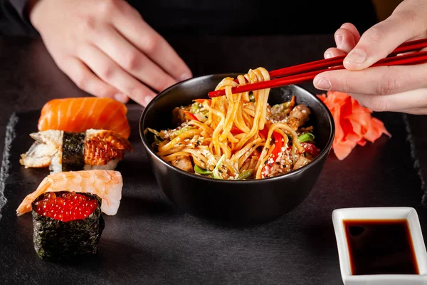 Concept of Asian cuisine. The girl is holding Japanese chopsticks in her hand and eating Chinese noodles from a black plate in a restaurant. background image. copy space