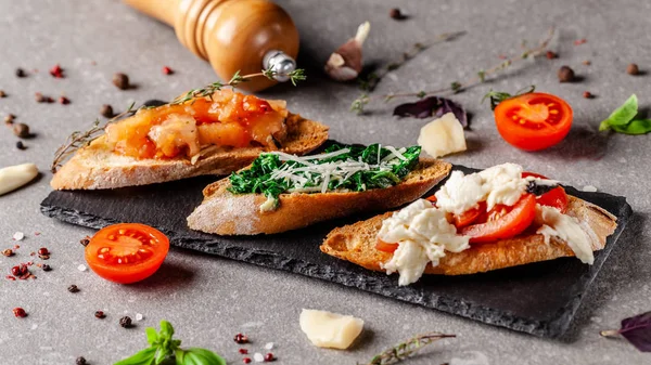 Concept of Spanish cuisine. Tapas Different bruschetta on a fried baguette with basil, tomatoes, cheese. Serving dishes in the restaurant. on a black stone slate board. copy space