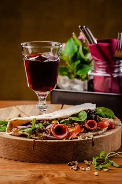 Koncept italská kuchyně. Piadina se šunkou, rajčaty, směsí salátu, pistácií, okurky na dřevěné desce. Sklenice červeného vína na stole. Nádherná jídla v restauraci. — Stock fotografie