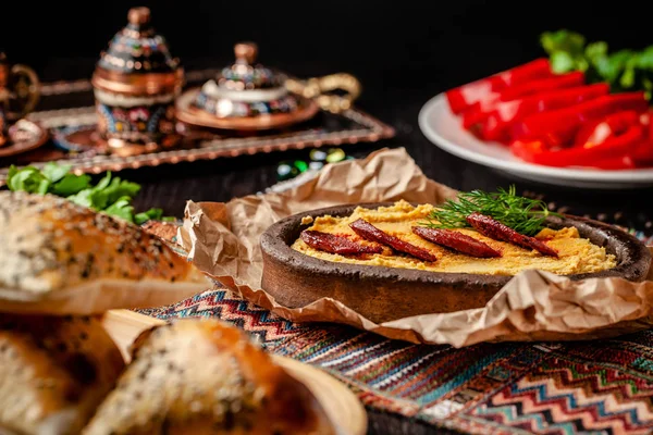 Традиционная турецкая, арабская кухня. Хумус с колбасой из салями, в глиняной тарелке, с пергаментом на деревянном столе. Подавать блюда в ресторане. копировальное пространство Стоковое Изображение
