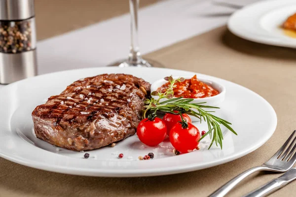 American cuisine. Beef steak with red bbq tomato sauce and cherry tomatoes. A glass of cool wine. Serving dishes on a white plate in a restaurant. Background image. copy space