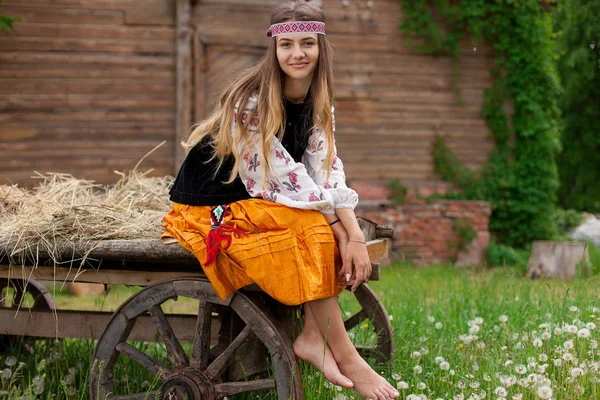 Beautiful slender Ukrainian woman in embroidery on the background of a wooden frame of a barn in the village sits on a wooden cart