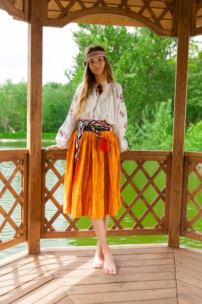 Beautiful Ukrainian woman dressed in embroidery in a wooden summer altanka on a lake