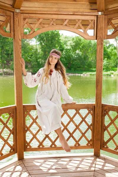 Beautiful Ukrainian woman dressed in embroidery in a wooden summer altanka on a lake