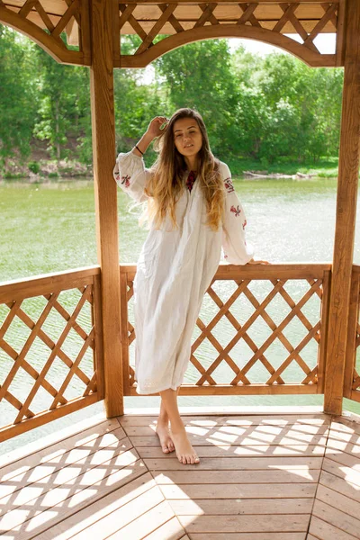 Beautiful Ukrainian woman dressed in embroidery in a wooden summer altanka on a lake