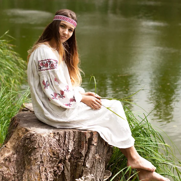 A beautiful Ukrainian woman dressed in embroidery sits on a stump on the shore of a lake