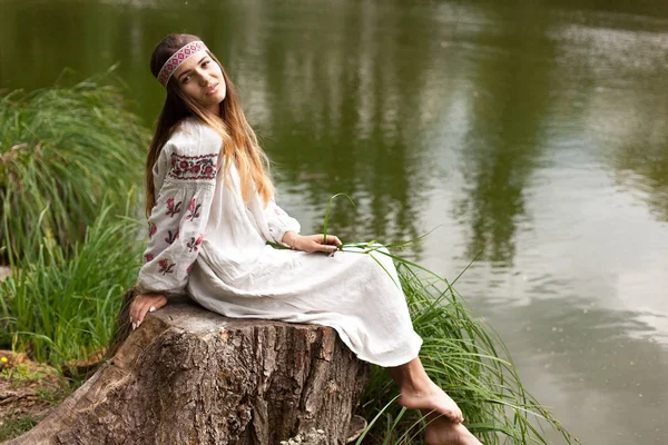 A beautiful Ukrainian woman dressed in embroidery sits on a stump on the shore of a lake