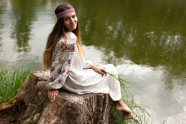 A beautiful Ukrainian woman dressed in embroidery sits on a stump on the shore of a lake