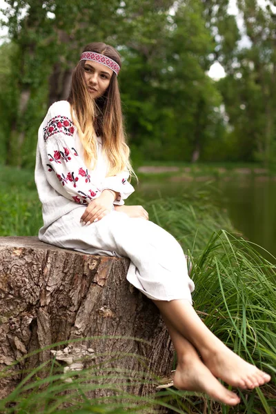 A beautiful Ukrainian woman dressed in embroidery sits on a stump on the shore of a lake