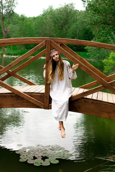 Beautiful Ukrainian woman wearing embroidery sits on a wooden bridge on a pond