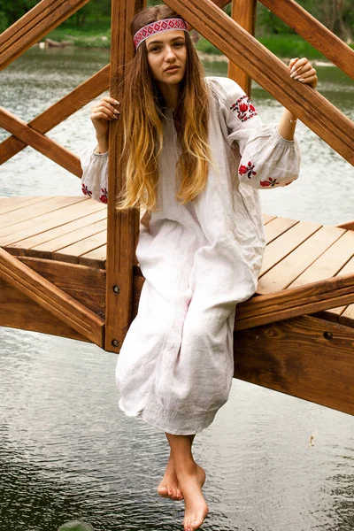 Beautiful Ukrainian woman wearing embroidery sits on a wooden bridge on a pond