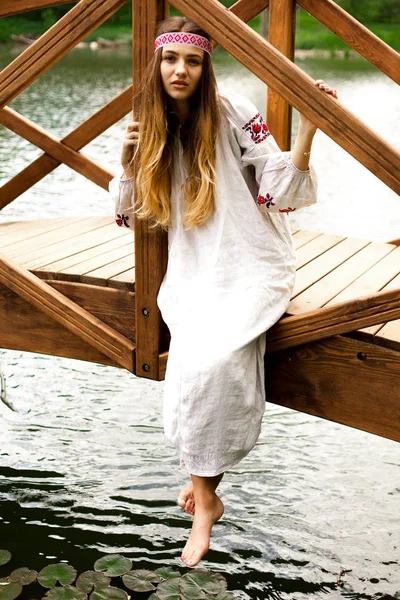 池に木製の橋に座っている刺繍を身に着けている美しいウクライナの女性 — ストック写真