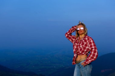 Kırmızı ekose gömlekli kız mavi sisli bir dağın arka planında poz veriyor.