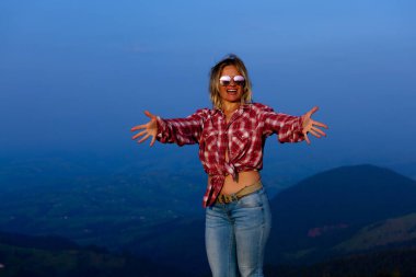 Kırmızı ekose gömlekli kız mavi sisli bir dağın arka planında poz veriyor.