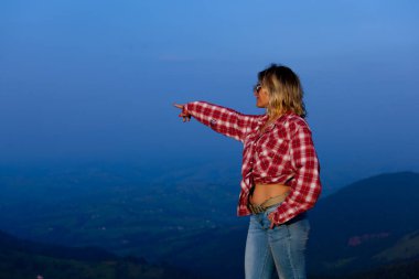 Kırmızı ekose gömlekli kız mavi sisli bir dağın arka planında poz veriyor.