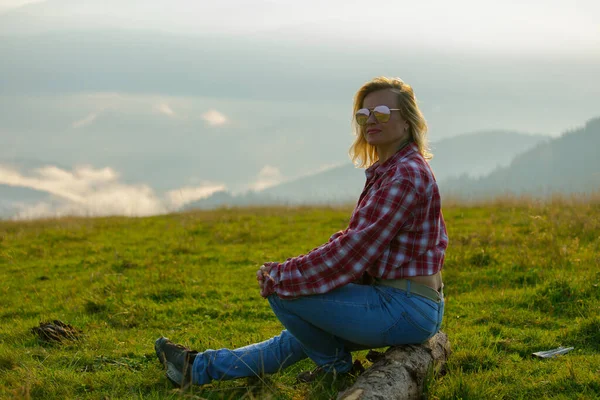 Blondynka Dżinsowych Ubraniach Okularach Siedzi Łące Mglistych Górach — Zdjęcie stockowe
