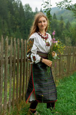 Bir buket kır çiçeğiyle gevşek saçlı kız tahta bir çitin önünde duruyor.