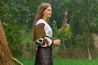 Bir buket kır çiçeğiyle dağınık saçlı kız samanlığın yanında duruyor.