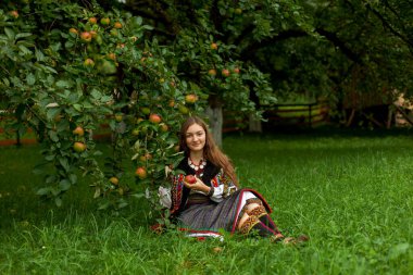 Nakış işlemeli olgun elmalı bir kız elma ağacının altındaki yeşil çimlerde oturuyor.