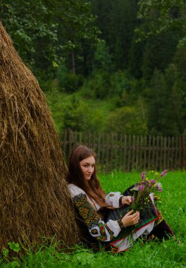 Bir buket kır çiçeğiyle dağınık saçlı bir kız sırtı samanlıkta oturuyor.