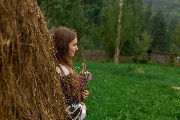 野花の花束を持つ緩やかな髪の女の子は干し草の山のそばに立っています — ストック写真