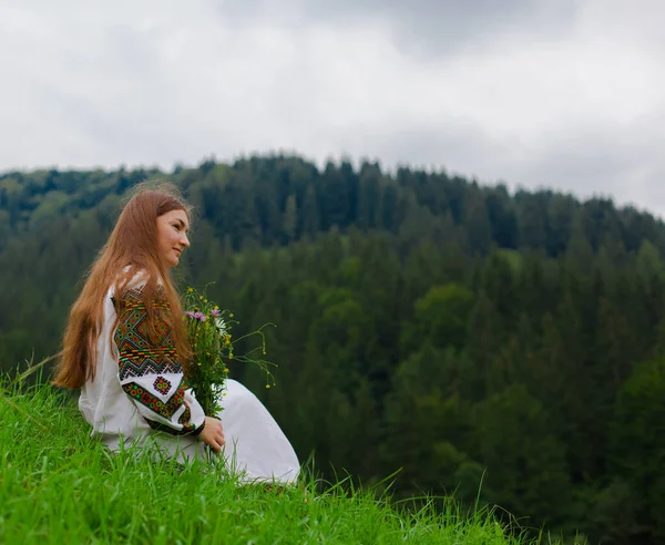 在喀尔巴阡山脉的背景上 坐在绿草上 她的头发蓬松 绣着一束野花 — 图库照片