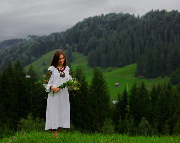 野生の花の花束を持つ刺繍の女の子はカルパチア山脈の緑の草の上に裸足で立っています — ストック写真