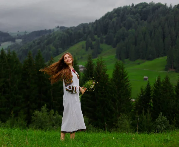 Bir buket kır çiçeğiyle nakış işlemeli gevşek saçlı kız Karpatya dağlarında mutlu bir şekilde koşar.
