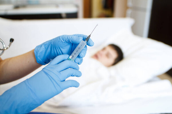 The doctor holds a syringe with a solution in the background of a bed-patient patient in the house