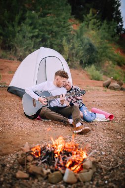 Bir adam ve bir kız kamp ateşi ve nehir tarafından oturmak ve gitar çalmak.