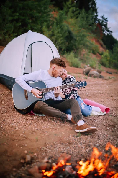 A man and a girl sit by the river and campfire and play the guitar. — Stock Photo, Image