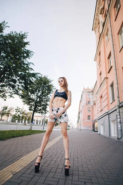 Menina de salto alto posando entre as ruas da cidade. Menina em um top preto e shorts curtos . — Fotografia de Stock