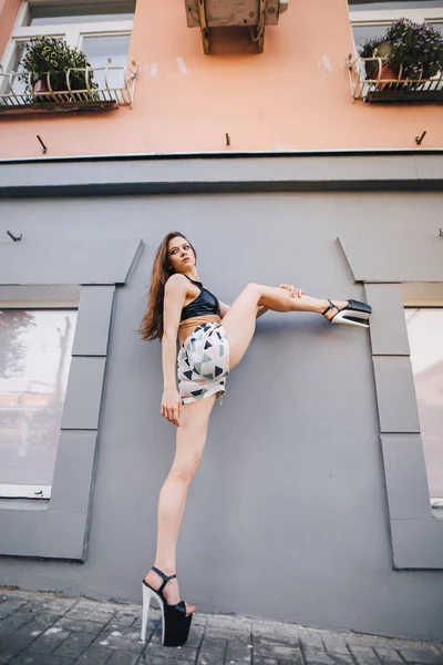 Una chica con tacones altos estira las piernas por la ventana de un edificio. Chica en un top negro y pantalones cortos Imagen de stock