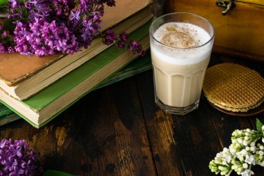 Lezzetli latte macchiato, bisküvi, eski kitaplar ve ahşap bir masa üzerinde leylak çiçekler
