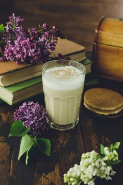 Lezzetli latte macchiato, bisküvi, eski kitaplar ve ahşap bir masa üzerinde leylak çiçekler