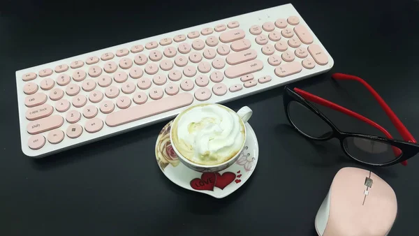Pink keyboard, mouse. up of coffee with cream. Feminine black and red eyeglasses. Black dark background. Saucer with flowers and love hearts.