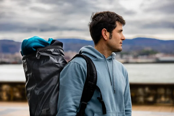 A side view of a young man in a hoodie carrying a travel backpack, looking out over the sea from a promenade.
