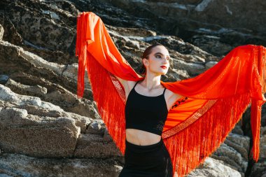 Young flamenco dancer posing with red shawl extended like wings, standing on rocky coastline during sunset, expressing grace and strength