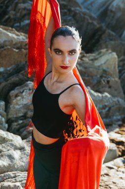Vertical portrait of a young flamenco dancer looking at camera, holding a red shawl over her shoulder, standing on a rocky coastal setting during sunset