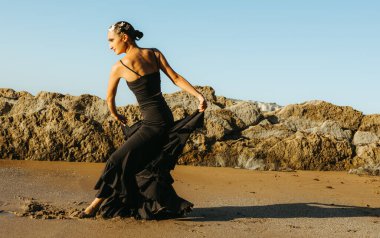 Young flamenco dancer in a black outfit dancing barefoot on the beach at sunset, gracefully posing near rocks with focus and passion