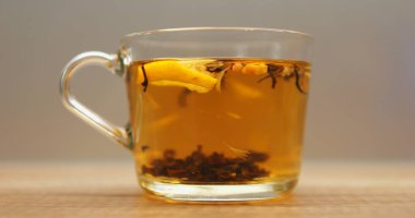 Çay yaprakları ve taze limon dilimleri şeffaf bardakta sıcak suyla döner. Ahşap masada cam bardakta bitki içeceği demleme işlemi