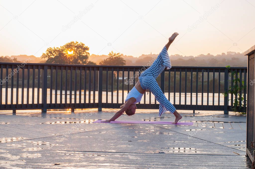 Young woman practicing yoga, standing in Downward facing dog exercise on the morning sunrise.