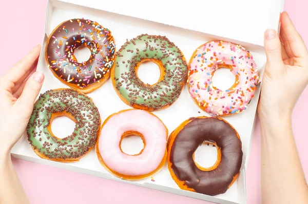 Female hands holding box with colorful donuts on pastel pink background