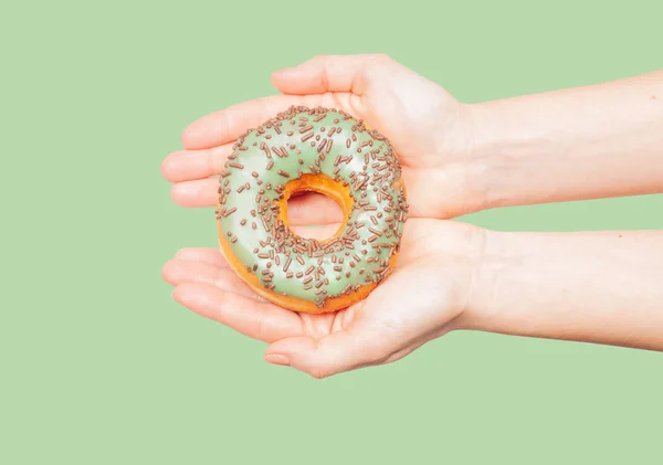 Manos femeninas sosteniendo rosquilla colorida sobre fondo verde pastel — Foto de Stock