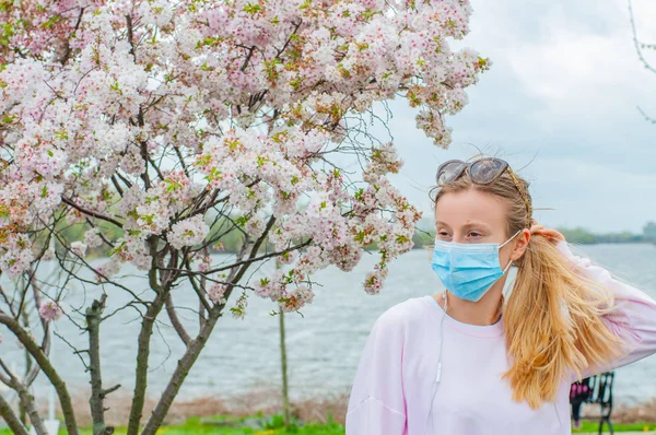 Alergia. Mulher nova na máscara protetora da alergia do pólen, entre árvores florescendo no parque . — Fotografia de Stock
