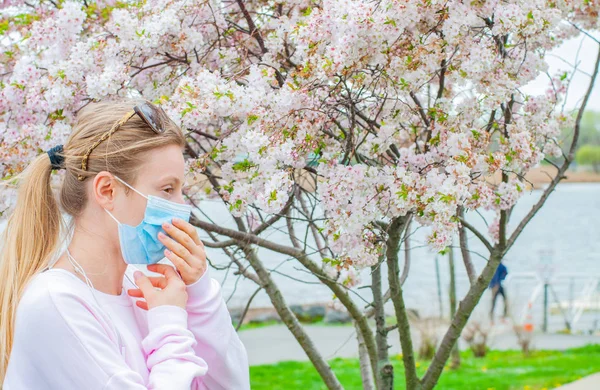 Alergia. Mulher nova na máscara protetora da alergia do pólen, entre árvores florescendo no parque . — Fotografia de Stock