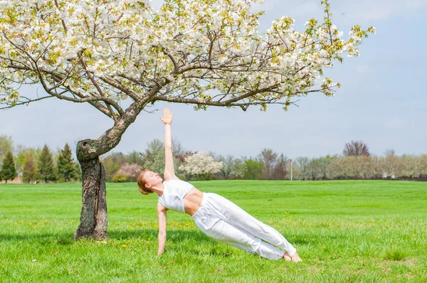 Hermosa mujer está practicando yoga, haciendo Camatkarasana pose cerca del árbol de flores — Foto de Stock