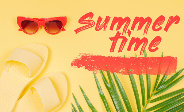 Summer Time text on yellow background, sandals and sunglasses with green tropical palm leaf.