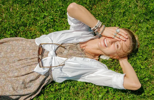 Красивая женщина в стиле бохо с большим количеством аксессуаров лежит на зеленой траве. Счастливая мамочка — стоковое фото